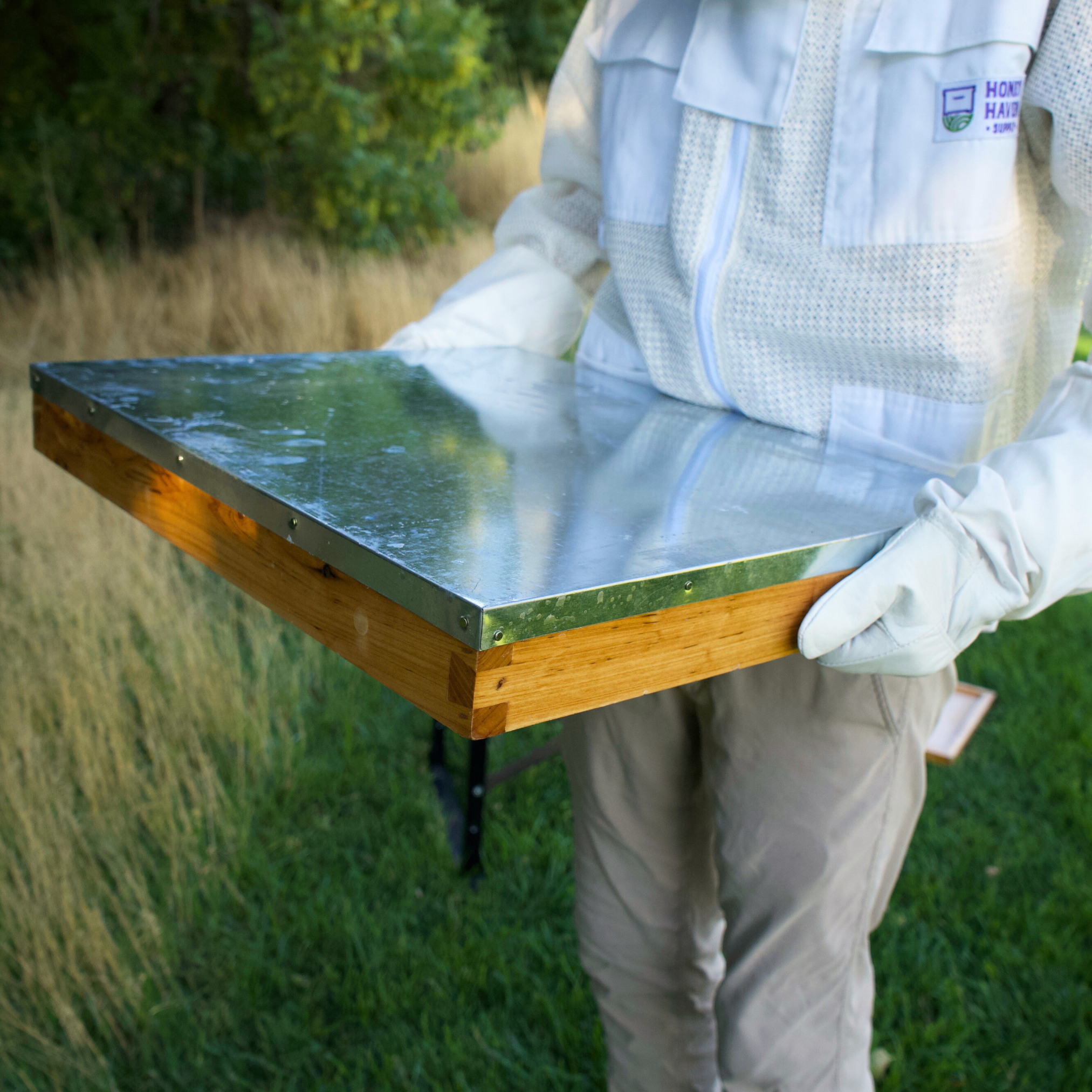 Man in a Honey Haven Supply branded ventilated bee suit carries a wax dipped beehive lid.