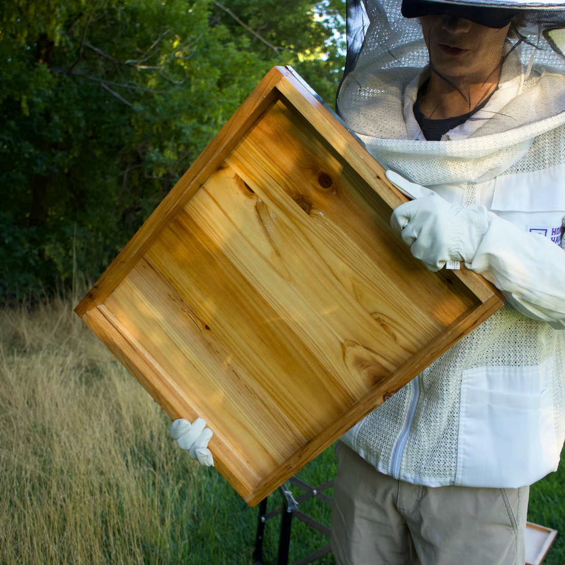 Man in a Honey Haven Supply branded ventilated bee suit carries a wax dipped beehive lid and holds it up to display the wax dipped underside.