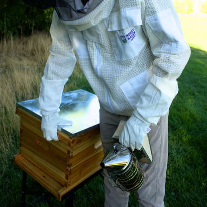 Man in white mesh ventilated beekeeping jacket using a smoker while standing by a double waxed hives.