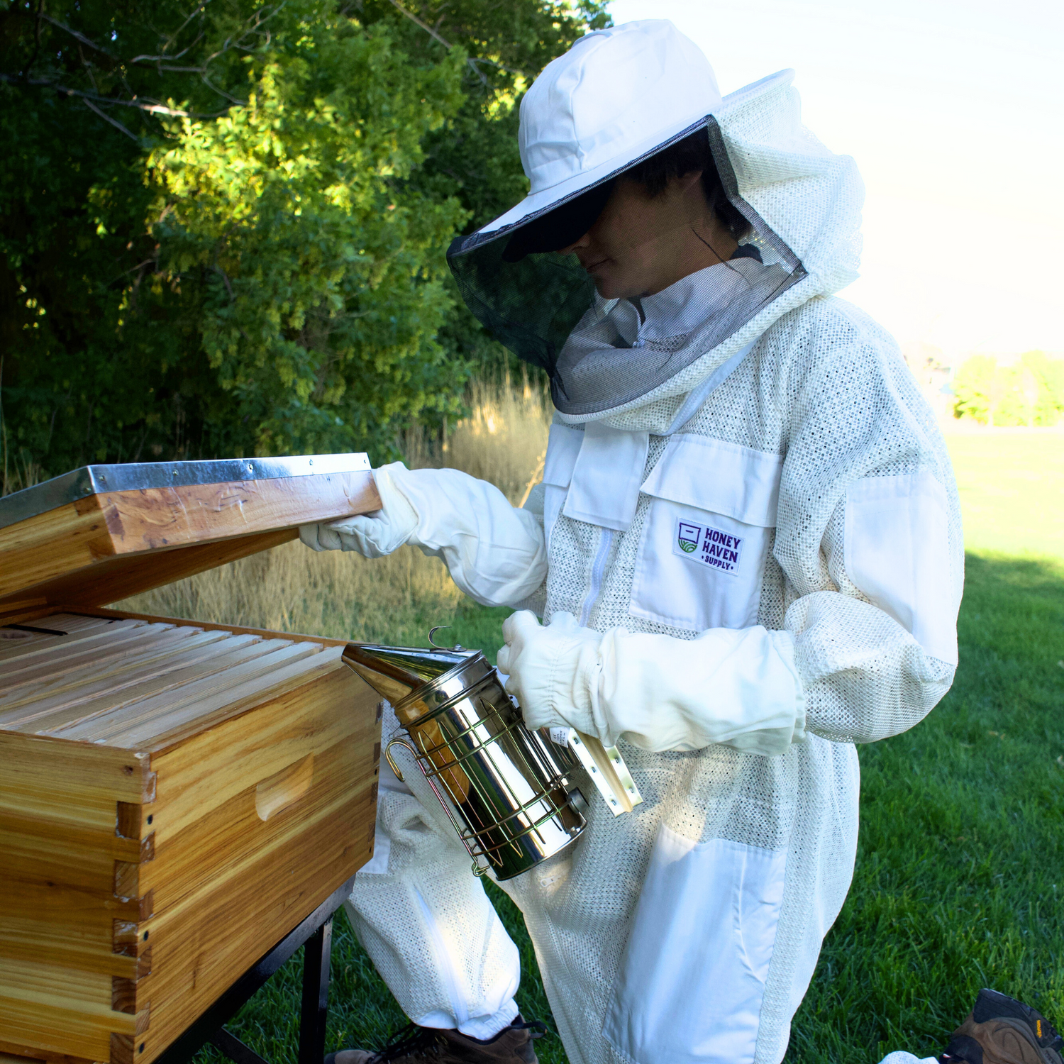 Man in white mesh ventilated beekeeping suit using a smoker while opening a double wax dipped hive