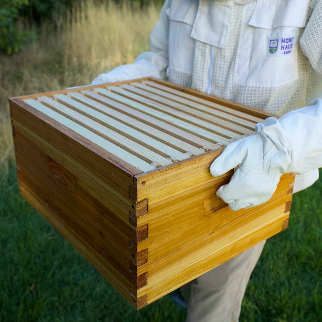 A man in a white ventilated beesuit holding a wax dipped deep brood box with frames.