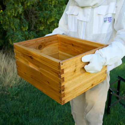 A man in a ventilated beekeeping suit holds a wax dipped deep brood box without frames in it.