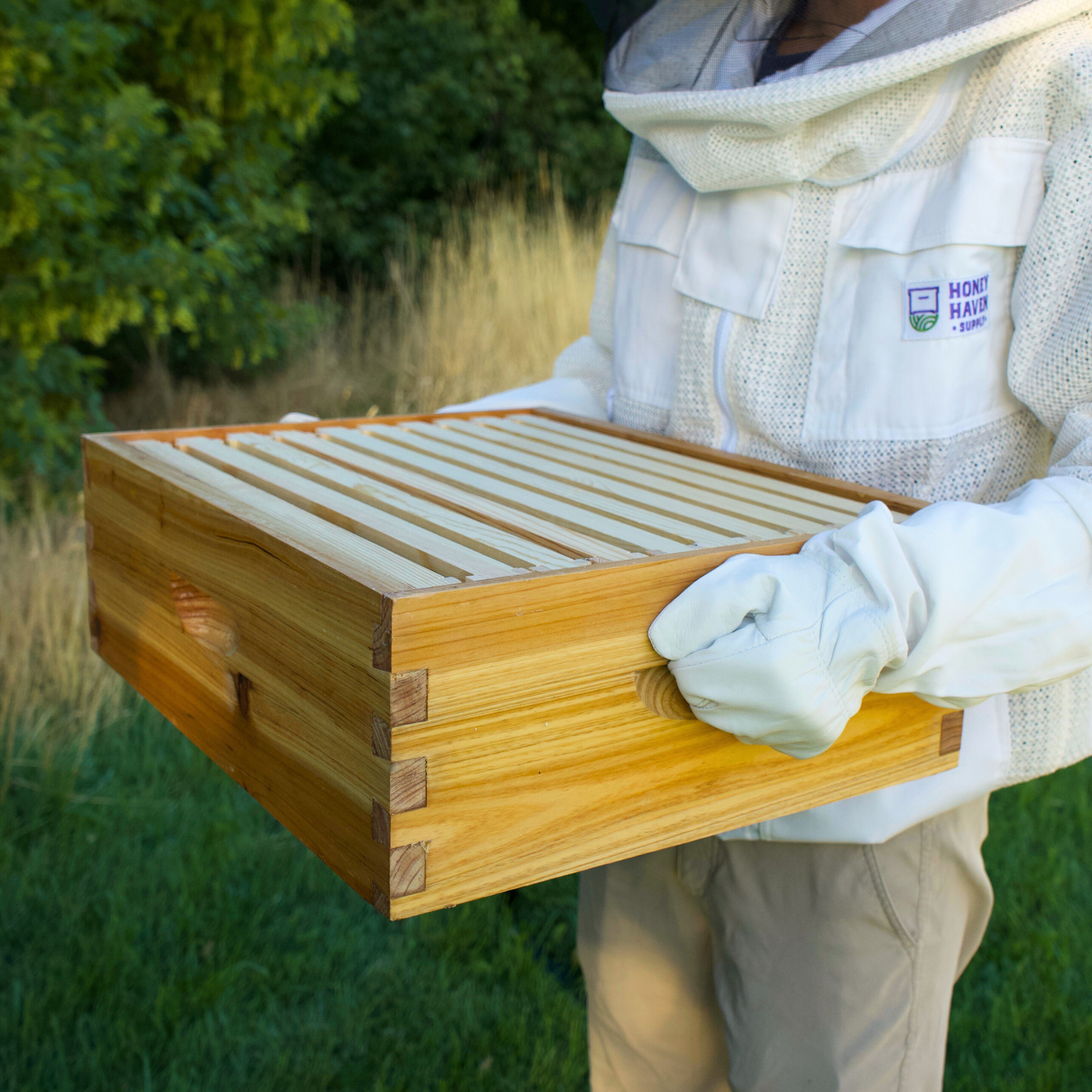 Man in beekeeping jacket holds a wax dipped honey super with frames in it.