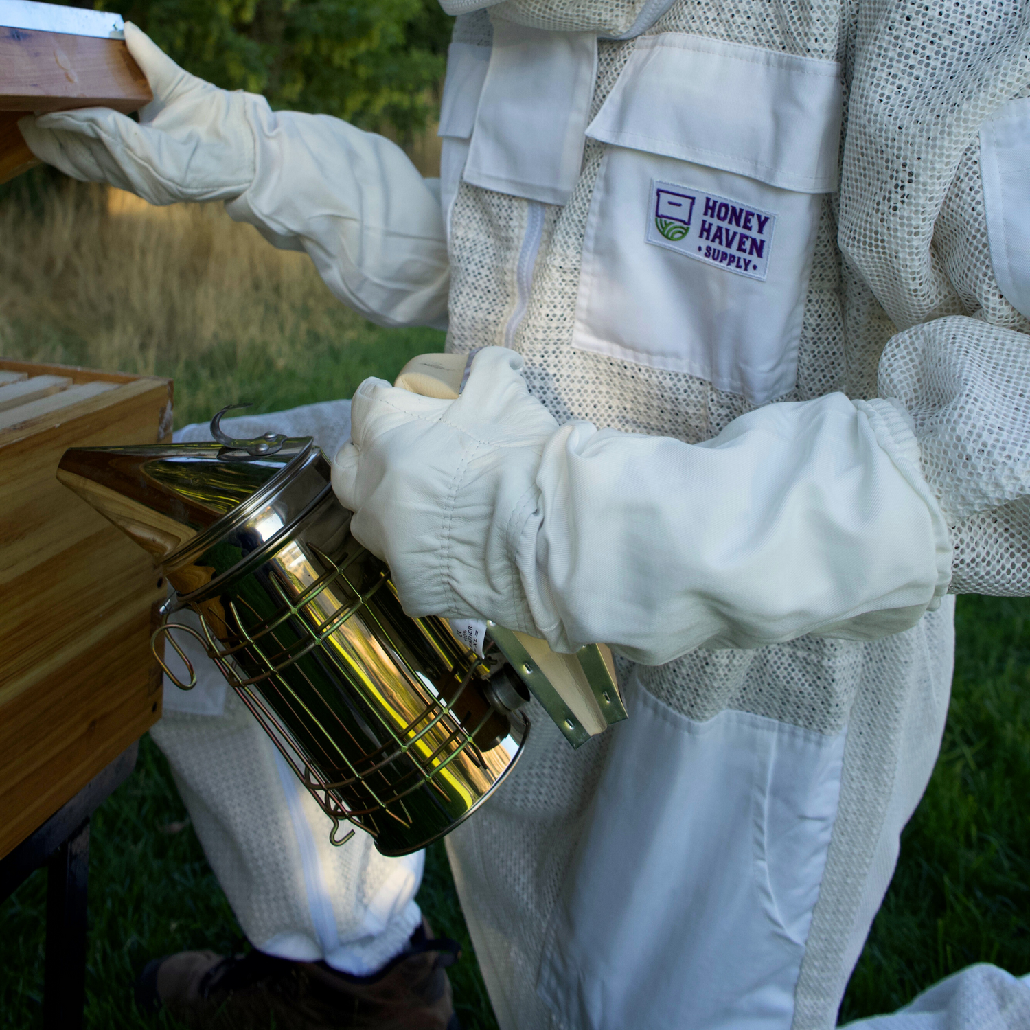 Close up of a man in a ventilated bee suit holding a smoker with white leather beekeeping gloves on.