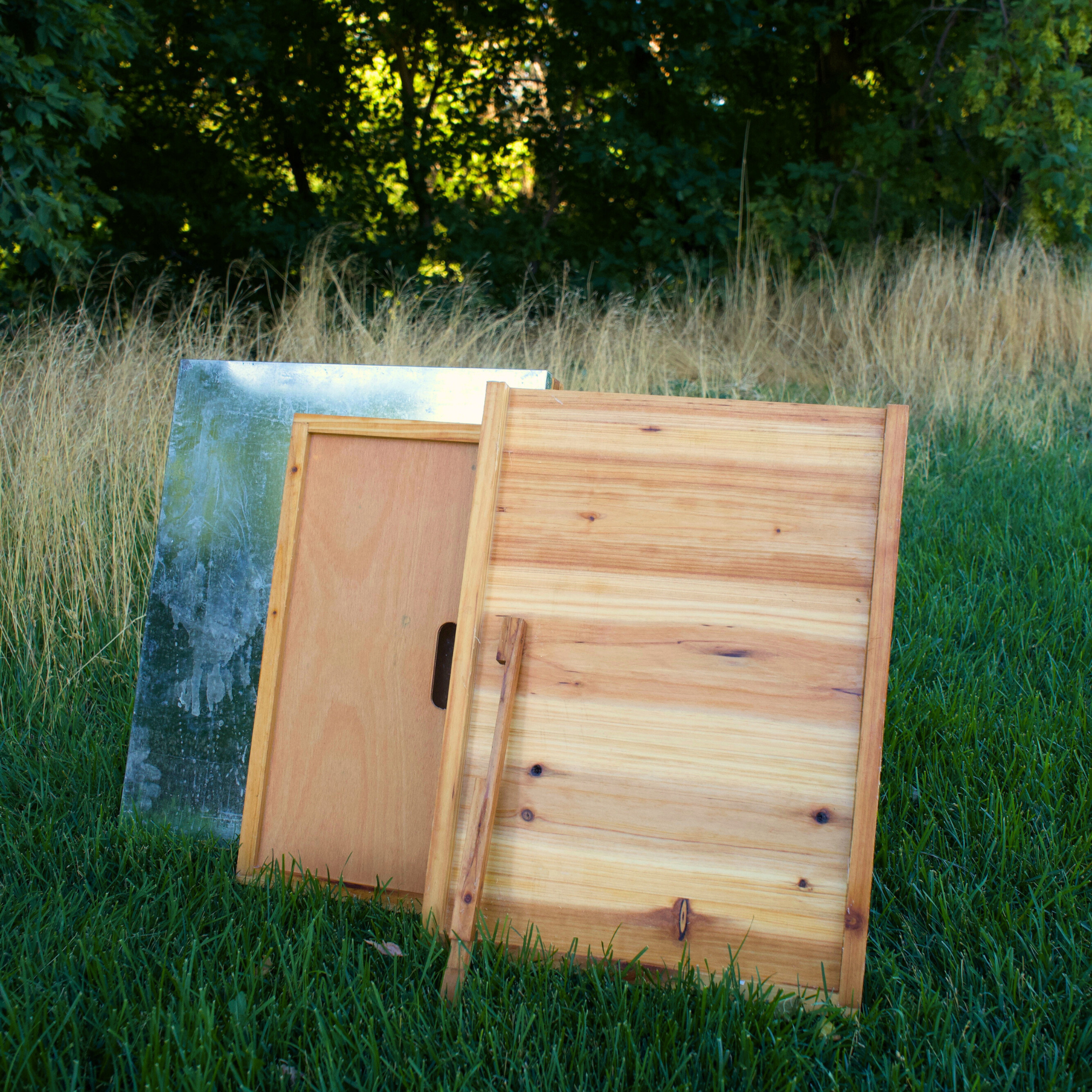 A beehive lid, inner cover, bottom board, and entrance reducer are all displayed in a green grass field. All these components are made from high quality pine and are double wax dipped!