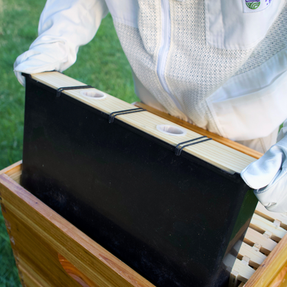 Man in bees suit displaying a internal plastic beehive feeder by pulling it halfway out of a hive.