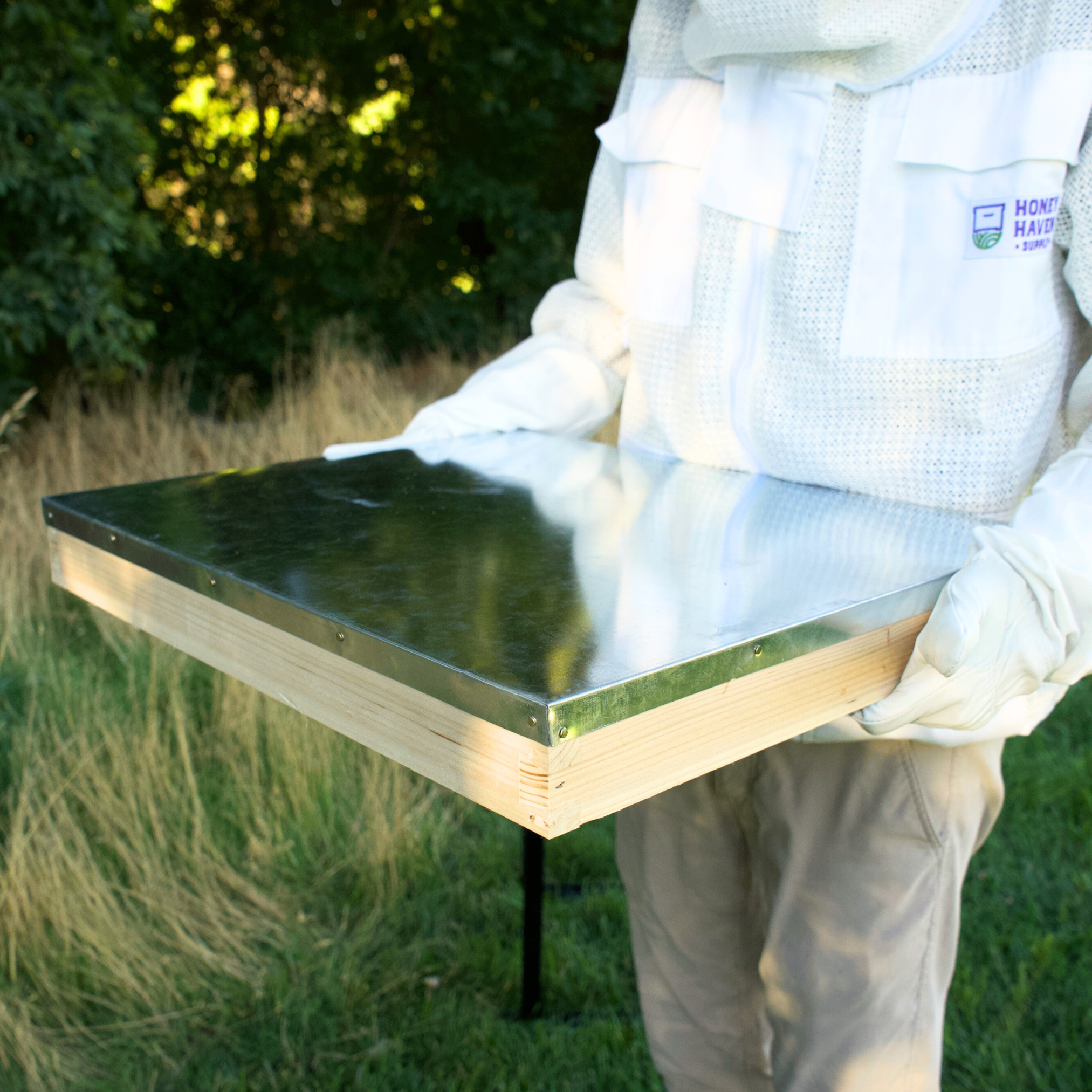 Man in white ventilated bee suit holds an untreated langstroth beekeeping lid.